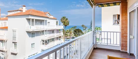A balcony with a wooden bench, overlooking a coastal area with buildings and palm trees.