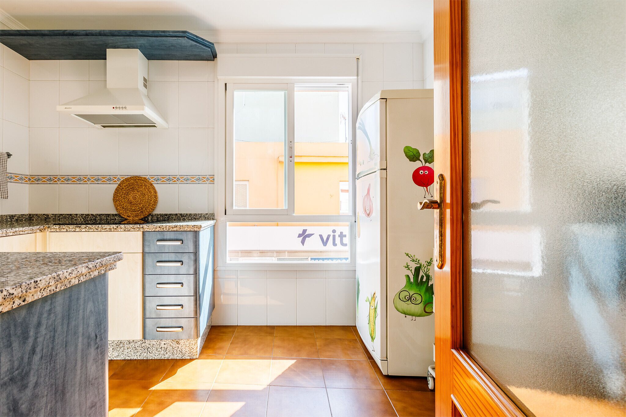 A kitchen with a window, a refrigerator with vegetable stickers, and a countertop with a woven basket.