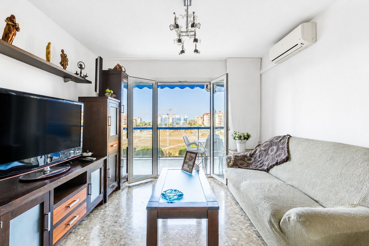 A modern living room with a flat-screen TV, a sofa, a coffee table, and a balcony with a view of buildings and a construction site.
