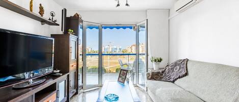 A modern living room with a flat-screen TV, a sofa, a coffee table, and a balcony with a view of buildings and a construction site.