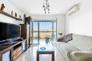 A modern living room with a flat-screen TV, a sofa, a coffee table, and a balcony with a view of buildings and a construction site.
