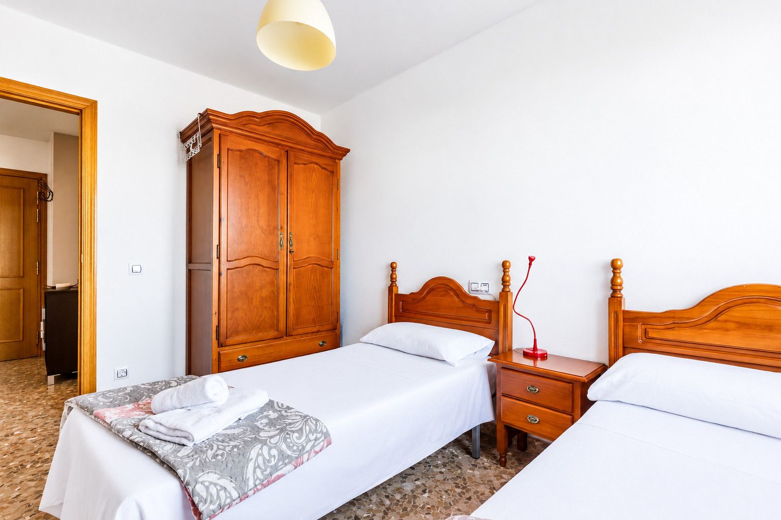Quarto de hotel com duas camas de solteiro, um guarda-roupa de madeira, mesa de cabeceira com abajur e piso ladrilhado.