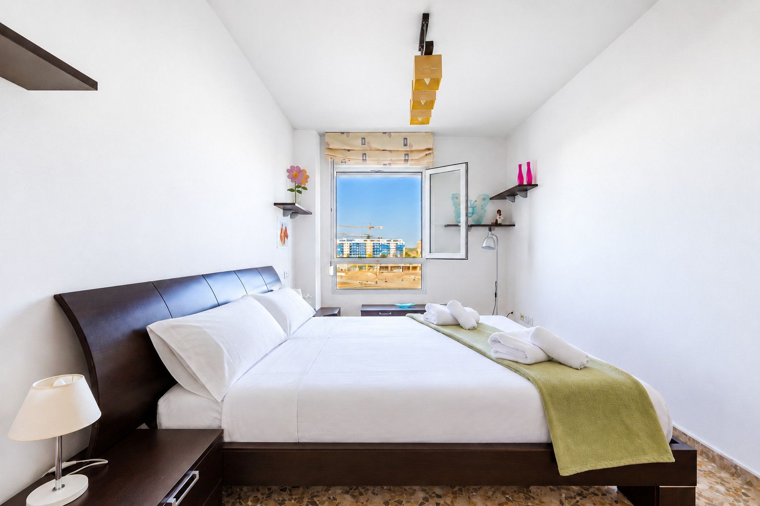 A hotel room with a bed, a nightstand, a lamp, a window with a view of buildings, and a shelf with decorative items.