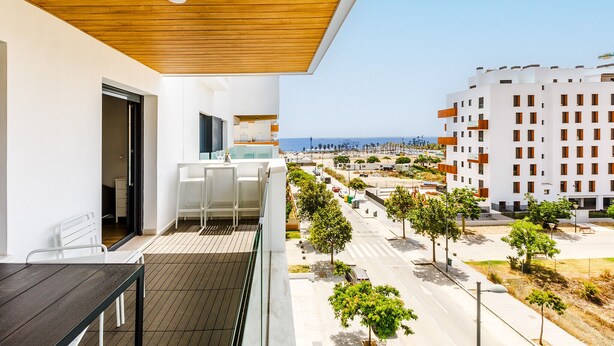 Un balcon doté d’un plafond en bois, de meubles blancs et offrant une vue sur une zone côtière avec des bâtiments et des arbres.