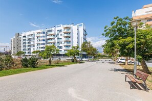 Ein modernes Mehrfamilienhaus mit mehreren Balkonen, einer gepflasterten Straße und einem Park mit Bänken.