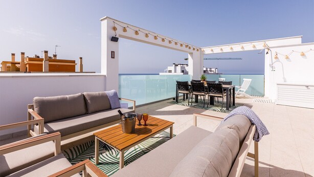 Une terrasse sur le toit aménagée avec un canapé, des chaises, une table et une pergola décorée de guirlandes lumineuses.