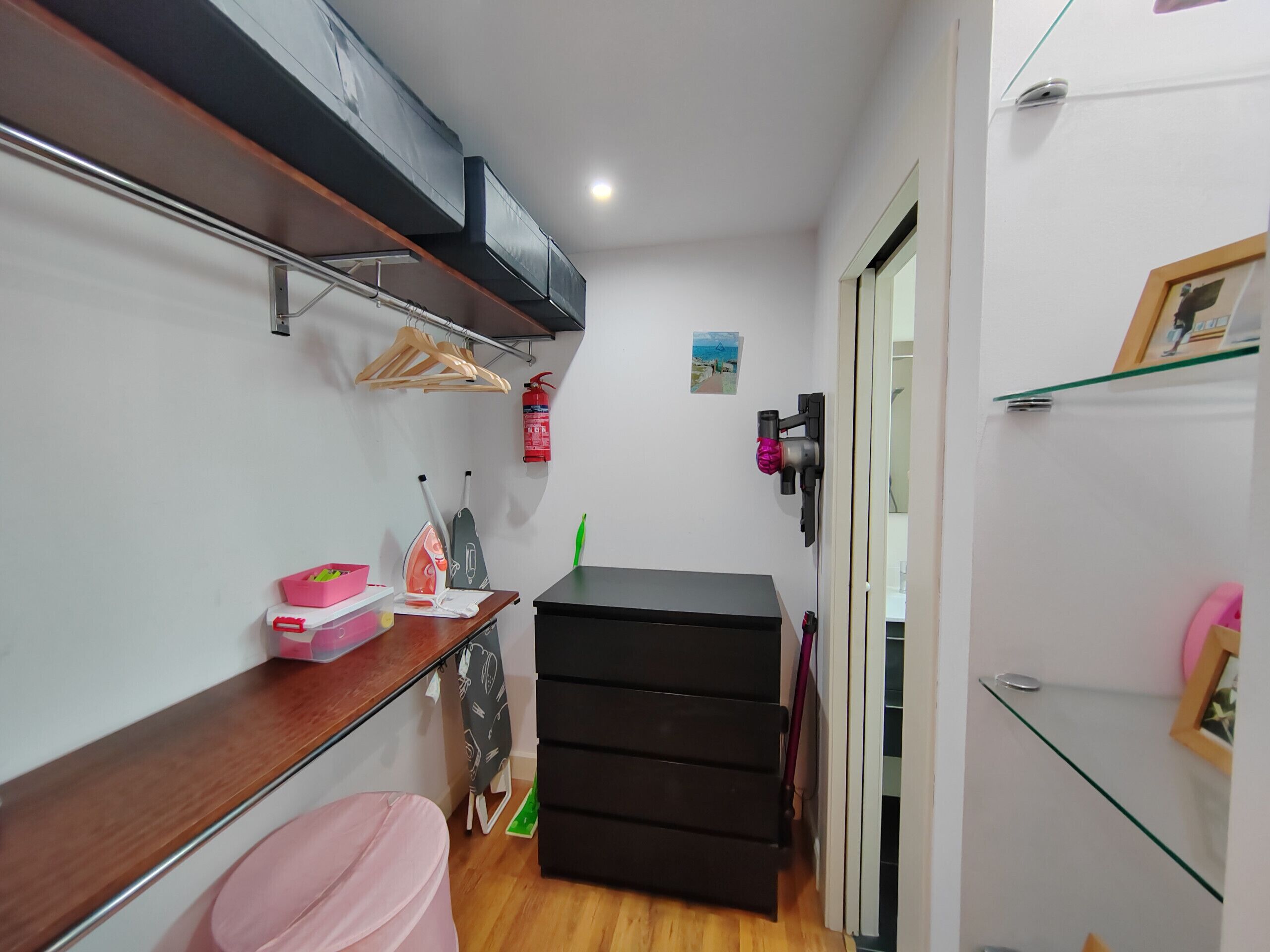 A compact room with a wooden bench, a black storage unit, a pink stool, and a small table with cleaning supplies.