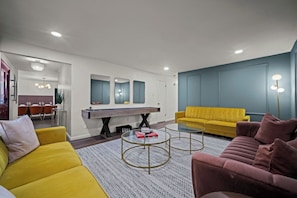 A modern living room with a yellow sofa, a pink sofa, a glass coffee table, and a dining area with a wooden table and chairs.
