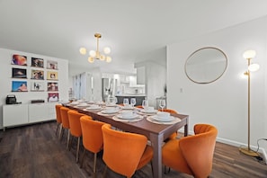 A dining area with a long table set for six, orange chairs, a chandelier, a round mirror, and a white cabinet with framed pictures.