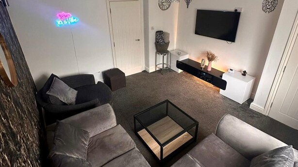 Un salon moderne avec un canapé d’angle gris, une table basse en verre, un meuble TV blanc et un panneau lumineux néon.