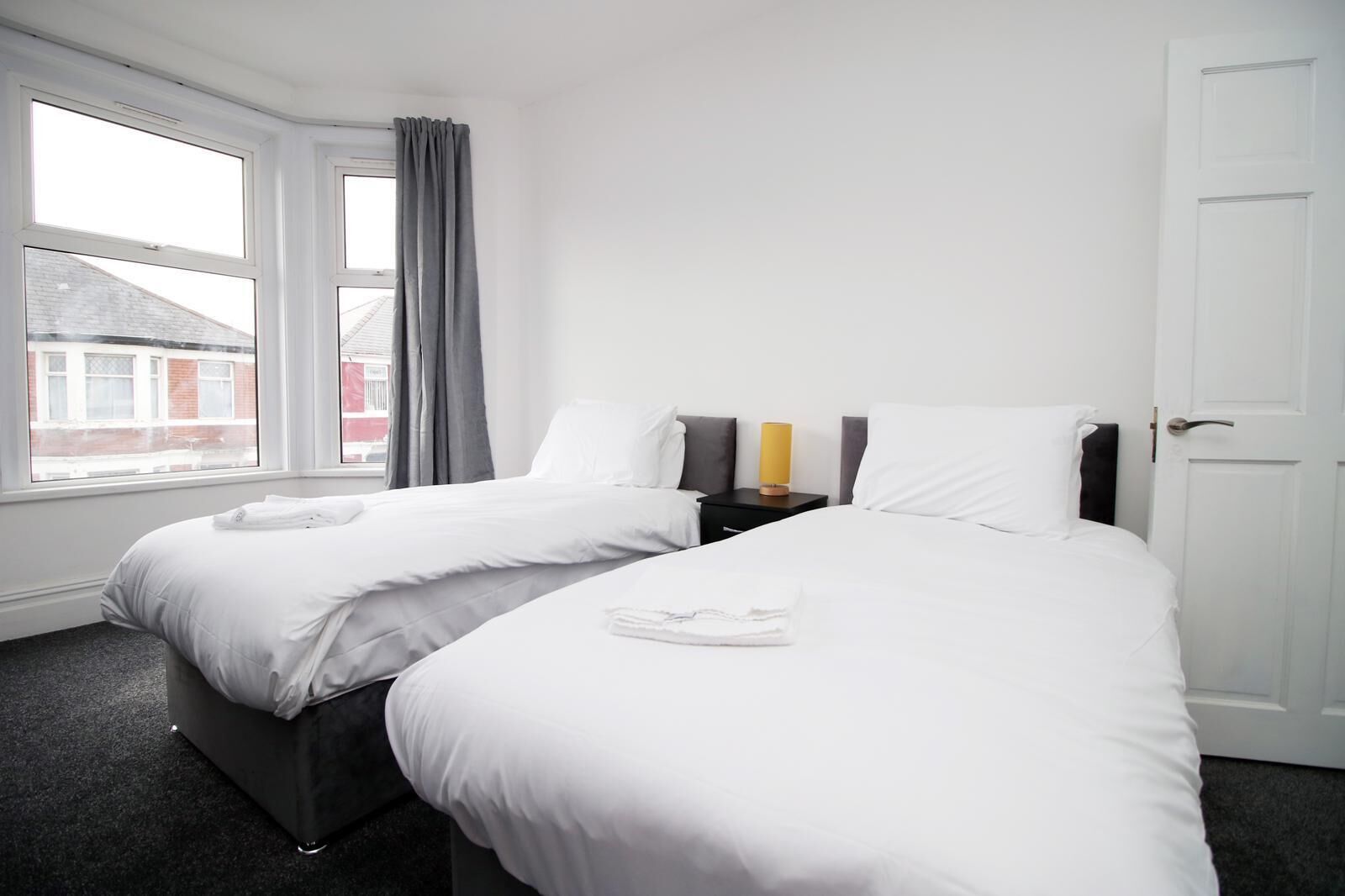 A hotel room with two beds, a window with curtains, a bedside table with a lamp, and a door.