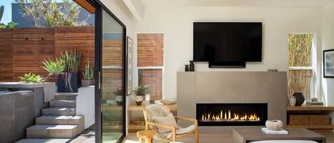 A modern living room with a fireplace, comfortable seating, a flat-screen TV, and a view of the outdoors.