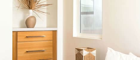 Un dormitorio con un aparador de madera, una mesita de noche con una lámpara decorativa, una ventana y una cama con ropa de cama blanca.