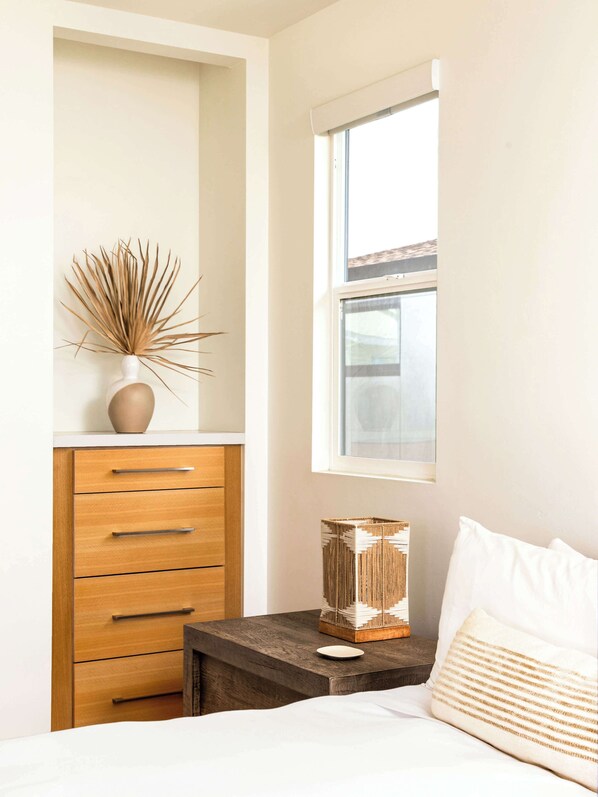 Un dormitorio con un aparador de madera, una mesita de noche con una lámpara decorativa, una ventana y una cama con ropa de cama blanca.
