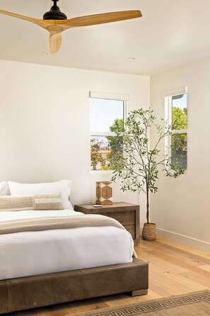 A bedroom with a bed, wooden floor, ceiling fan, windows, and a plant. - Sleek Beach House | Spa, Fire Pit & Ocean Views (Encinitas)