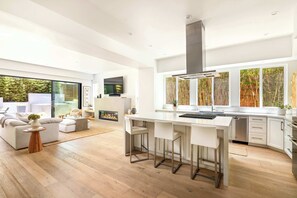 A modern kitchen with a central island, bar stools, and a large window with a view of trees. - Sleek Beach House | Spa, Fire Pit & Ocean Views (Encinitas)