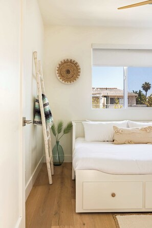 A bedroom with a white bed, a wooden ladder, a woven wall hanging, and a window with a view of buildings and palm trees.