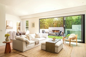 A modern living room with a sofa, coffee table, and chairs, a dining area with a table and chairs, and a large sliding glass door leading to a garden.