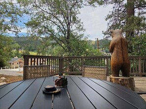Eine Holzterrasse mit einem Tisch, einer Bärenstatue und Blick auf ein bewaldetes Gebiet.