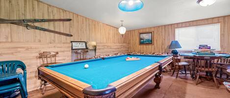 A pool table in a room with wooden walls, a blue ceiling light, and a window with blinds.