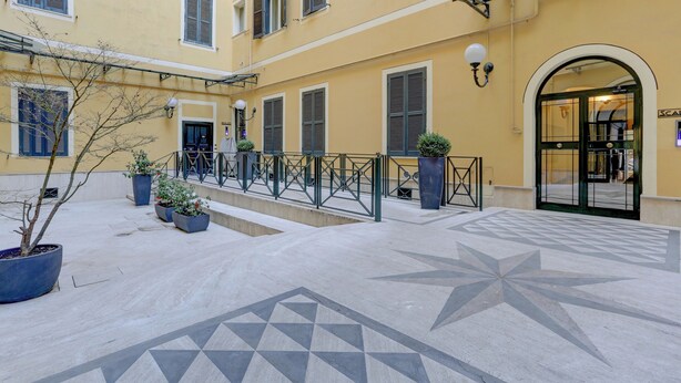 A courtyard with a geometric tile pattern, potted plants, a metal railing, and a building entrance with a glass door. - COLOSSEO Apt Mecenate 77 - Home and More (Rome)