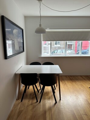 Ein Essbereich mit einem weißen Tisch und schwarzen Stühlen, einer Pendelleuchte, einem gerahmten Bild an der Wand und einem Fenster mit Blick auf Gebäude und Autos.