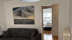 Ein modernes Wohnzimmer mit einem grauen Sofa, einem weißen Tisch und einem schwarzen Sessel. An der Wand hängt ein großes Gemälde und es gibt einen weißen Heizkörper.