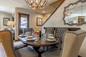 Un coin repas avec une table ronde dressée pour quatre personnes, entourée de chaises rembourrées dont les dossiers sont ornés de boutons. Un lustre est suspendu au plafond, et un miroir avec un cadre décoratif est fixé au mur.
