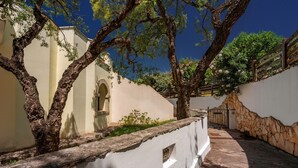 A courtyard with stone walls, a white building with an arched window, and trees with green foliage. - Beautiful Vista Blu Resort No2153 (Alghero)