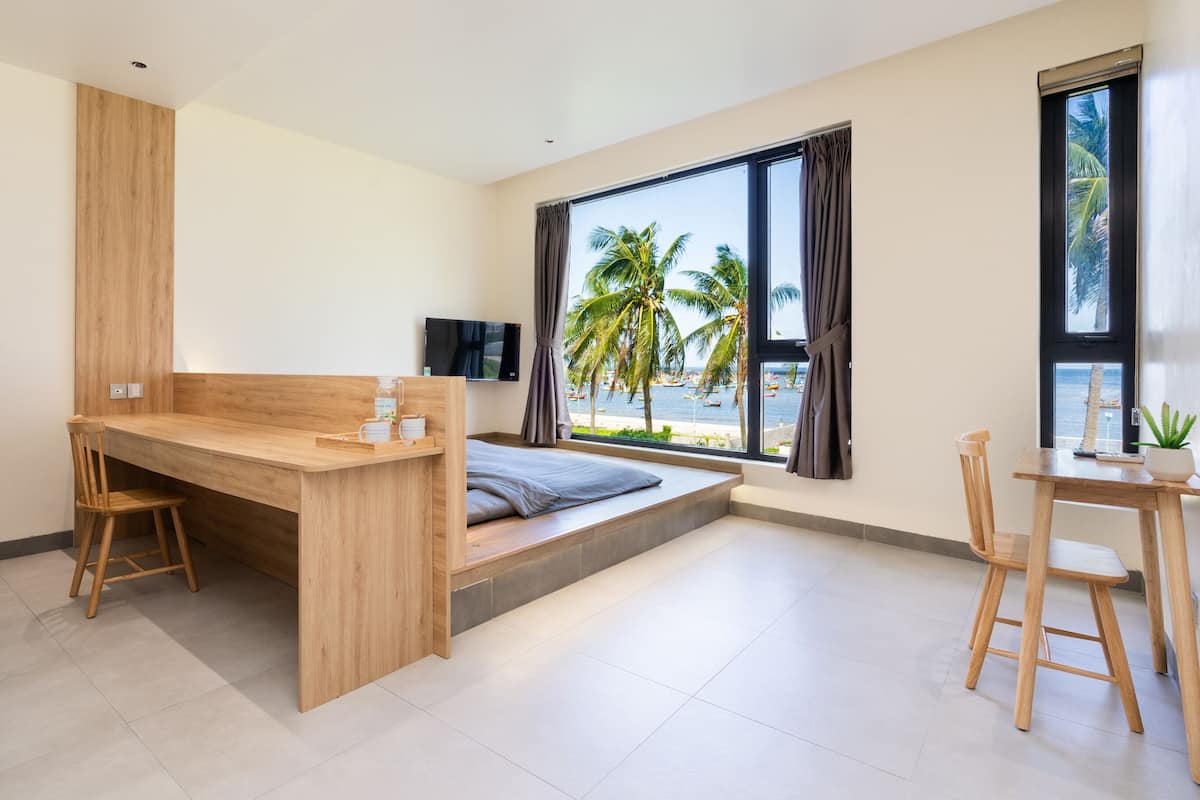 A modern room with a wooden desk, a bed with a view of palm trees, a TV, and a small table with a plant.