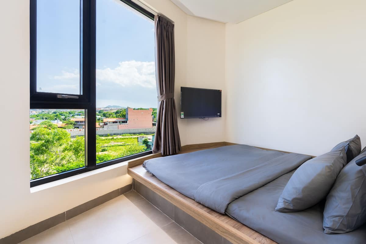 A bedroom with a large window, a flat-screen TV mounted on the wall, and a bed with grey bedding.