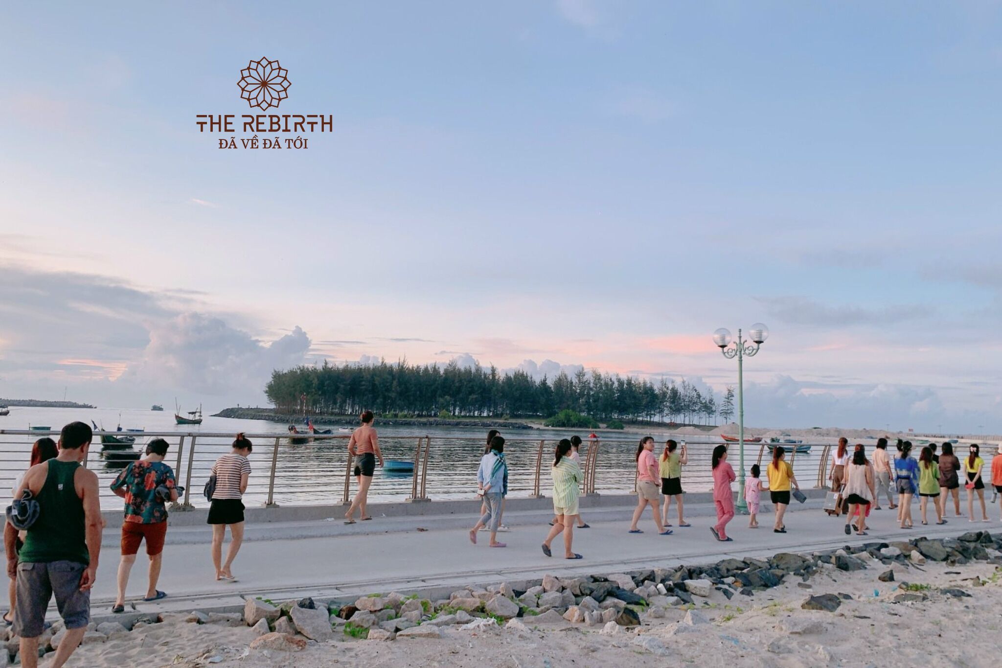 A waterfront promenade with people walking, a clear sky, and boats in the water.