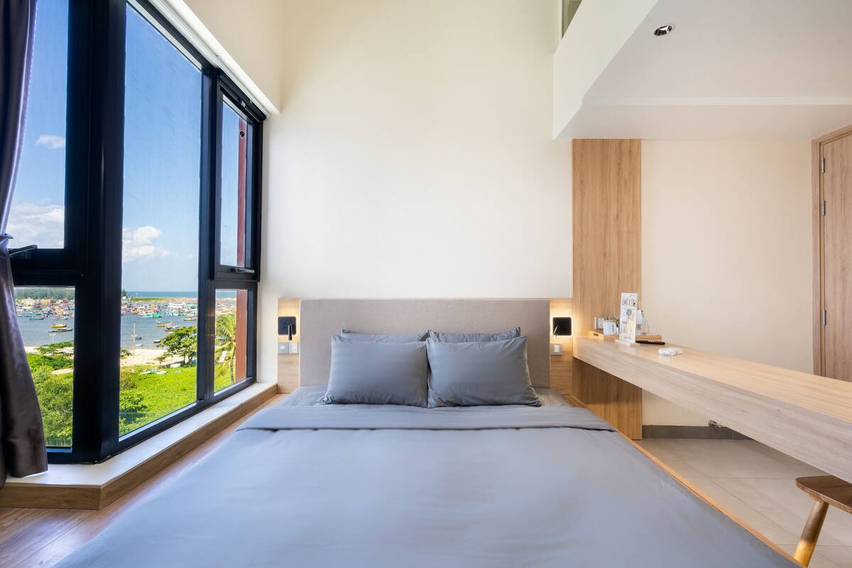 A modern bedroom with a large window, a bed with grey bedding, a wooden headboard, and a wooden bedside table with a lamp.