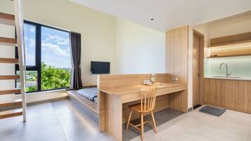 A modern room with a wooden desk, a chair, a bed, a TV, and a view of greenery outside.