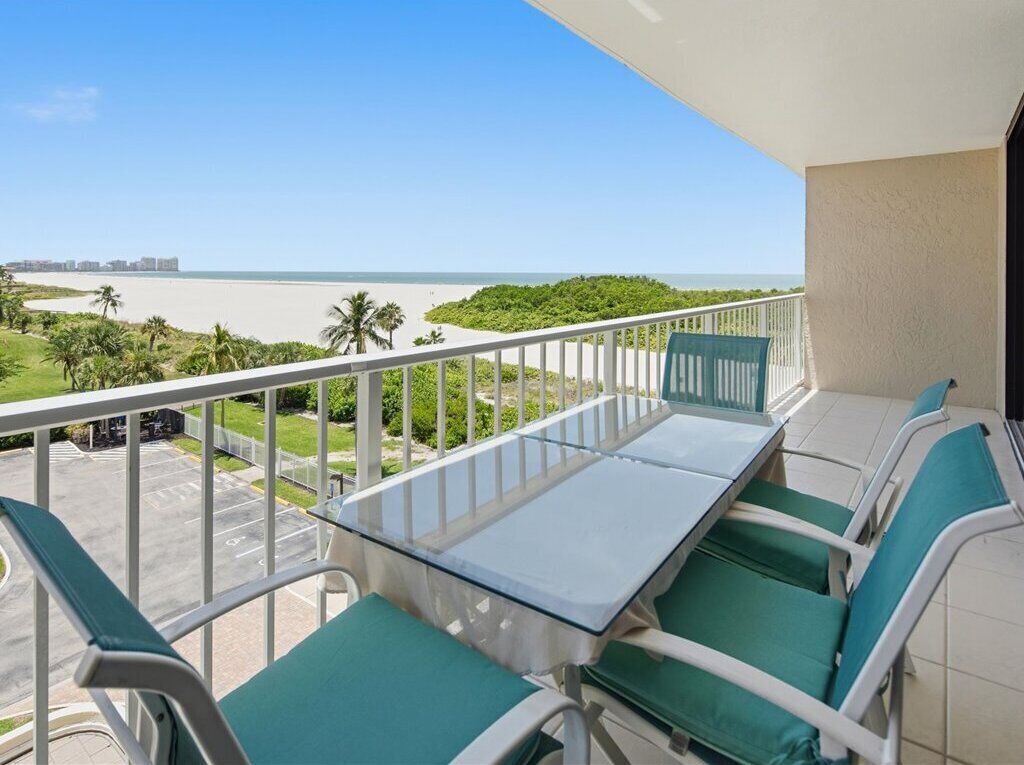 Varanda com mesa e cadeiras de vidro, com vista para uma praia e um estacionamento.