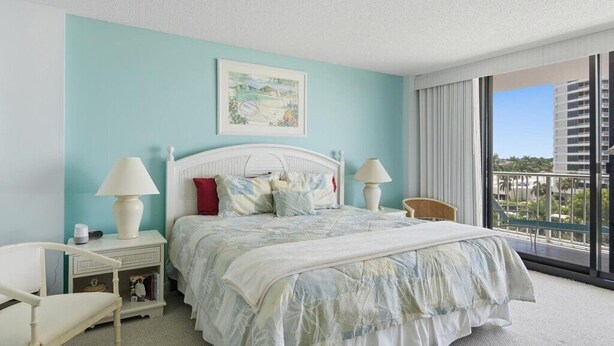 A bedroom with a large bed, two bedside tables, a chair, a window with a view, and a painting on the wall.