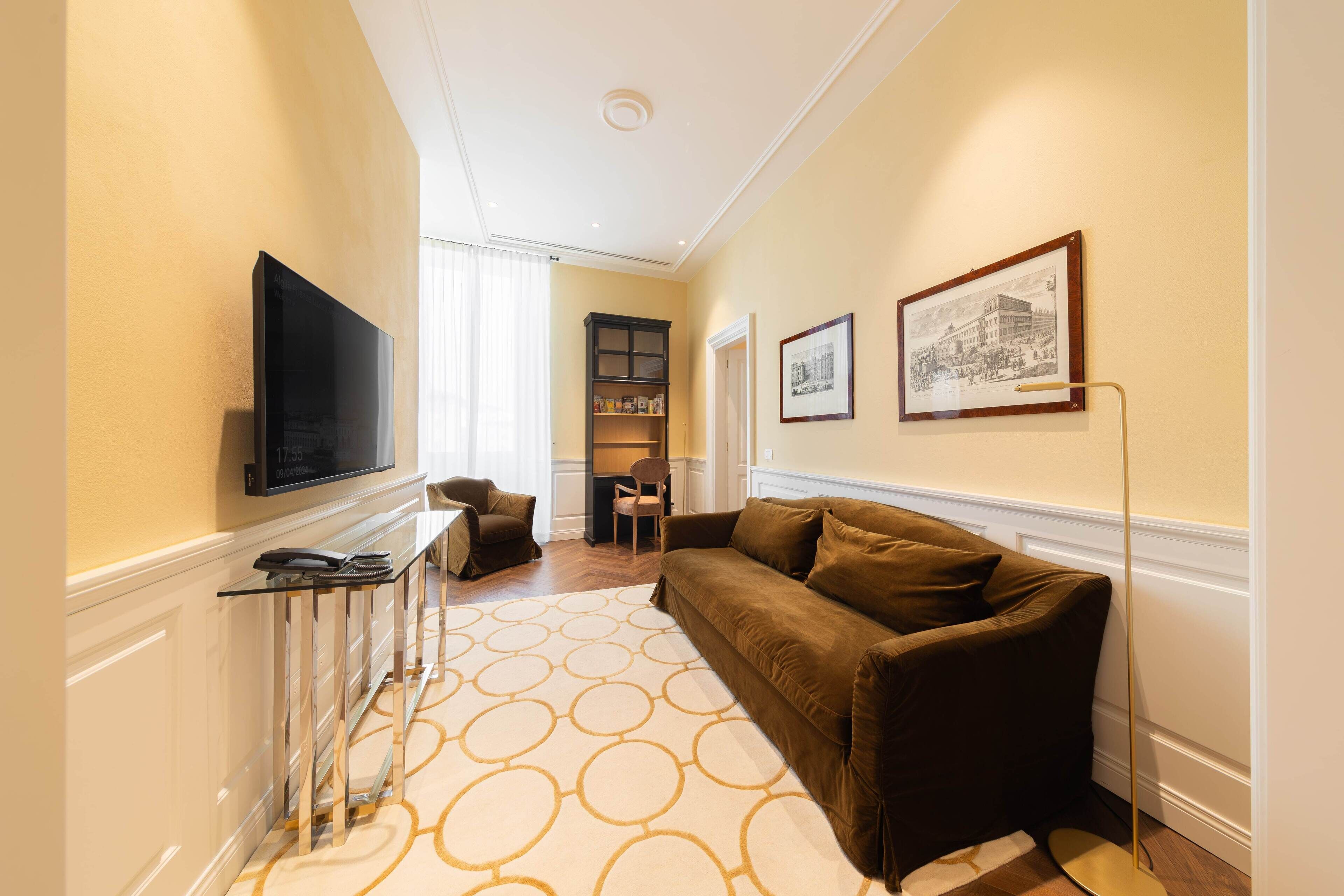 A modern living room with a brown sofa, a flat-screen TV, a glass coffee table, and a patterned rug.