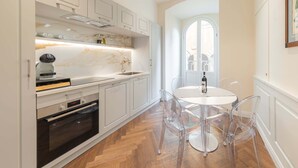 A modern kitchen with white cabinetry, a built-in oven, a marble backsplash, and a dining area with clear chairs and a white table. - Algilà Luxury Torrigiani Apartments (Florence)