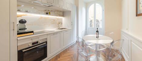 A modern kitchen with white cabinetry, a built-in oven, a marble backsplash, and a dining area with clear chairs and a white table.