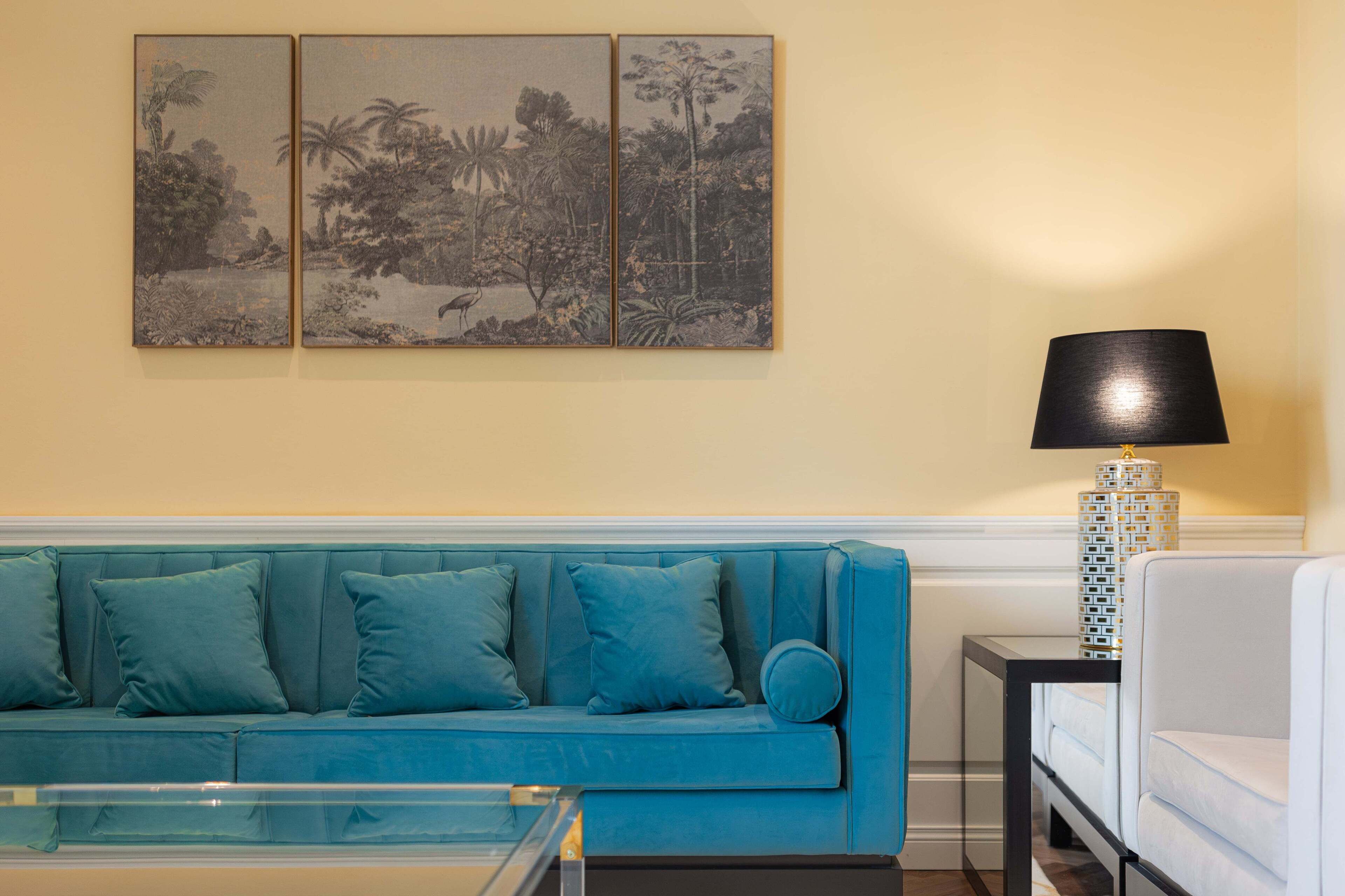 Un canapé de couleur bleu sarcelle agrémenté de coussins assortis, une table basse avec un plateau en verre, une lampe noire dont le pied est orné de motifs, et un triptyque décoratif fixé au mur.