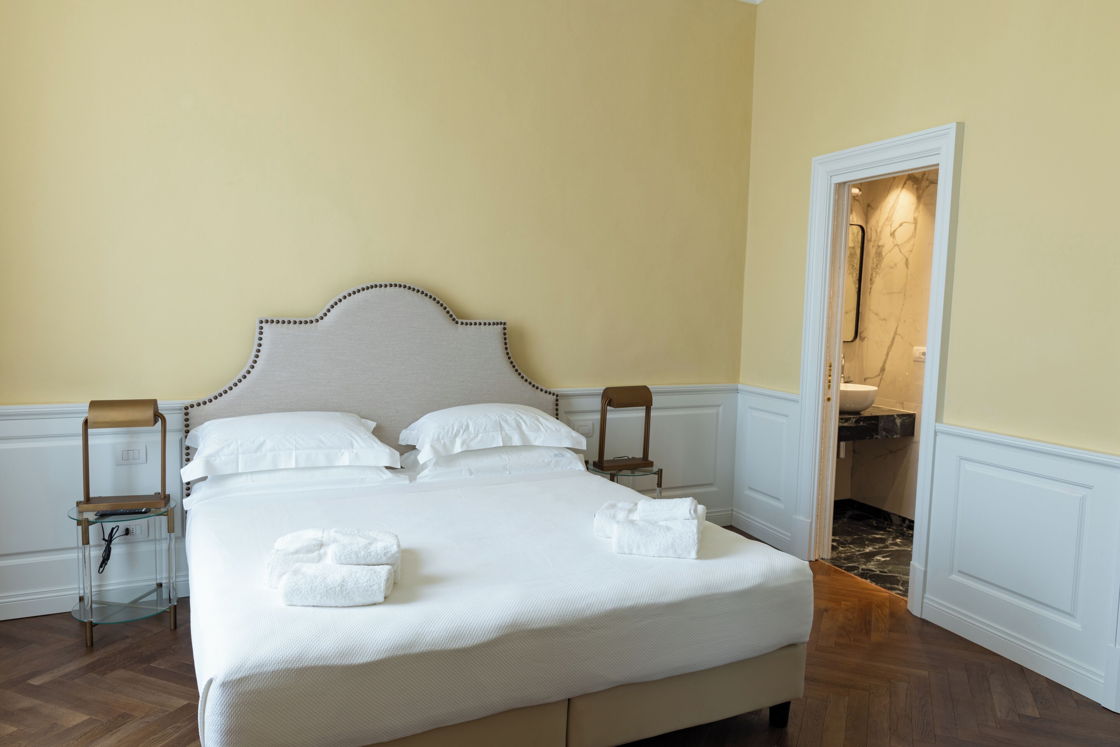 A neatly made bed with white linens and two towels, a wooden nightstand with a glass top, and a wooden chair beside the bed.
