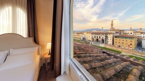 A hotel room with a large bed, a bedside table, and a view of historic buildings and a river. - Algilà Luxury Torrigiani Apartments (Florence)