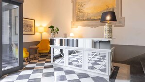 A lobby with a checkered floor, a white console table with a lamp, a painting on the wall, and a yellow chair. - Algilà Luxury Torrigiani Apartments (Florence)
