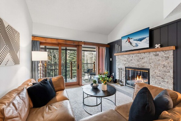 Un salon avec un canapé en cuir, une cheminée, un téléviseur et un balcon donnant sur la forêt.