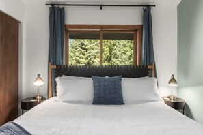 Une chambre à coucher avec un grand lit, deux lampes de chevet, une fenêtre avec des rideaux et une vue sur les arbres.
