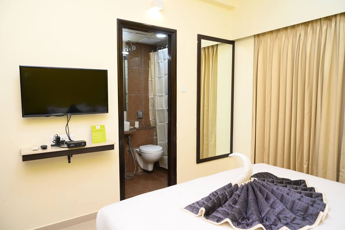 A hotel room with a bed, a TV mounted on the wall, a bathroom visible through an open door, and a mirror on the wall.