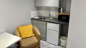 A compact kitchen with a sink, microwave, and a small refrigerator. There is a chair with a yellow cushion and a white table. - Beautiful 1-bed Modern Luxury Apartment in Luton (Luton)