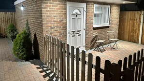 A small brick building with a white door and window, a wooden fence, and a patio with a chair and table. - Beautiful 1-bed Modern Luxury Apartment in Luton (Luton)