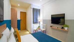 Een hotelkamer met een bed, een aan de muur bevestigde televisie, een klein tafeltje met stoelen en een deur naar een andere kamer.
