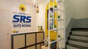 Een receptiebalie van een hotel met een bordje 'HOTEL SRS SUITE ROOMS', een trap en een plant aan de muur.